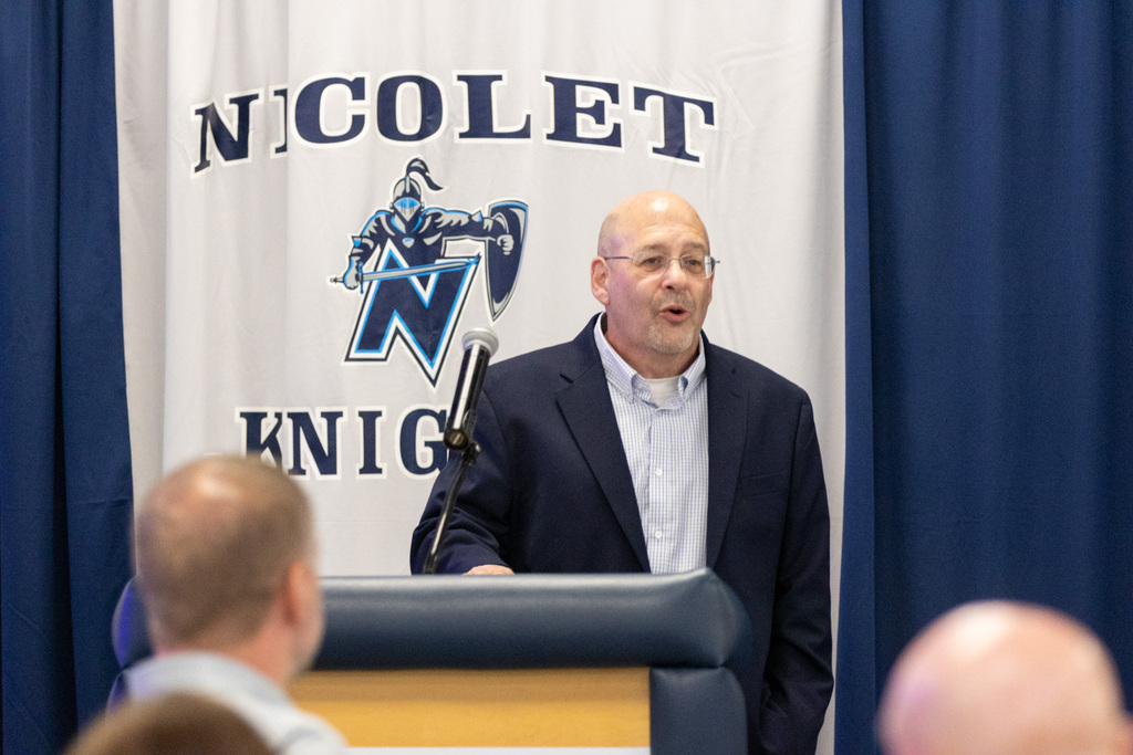 A man in glasses and a dark suit stands at a podium, speaking into a microphone. Behind him, a Nicolet Knights banner is displayed.
