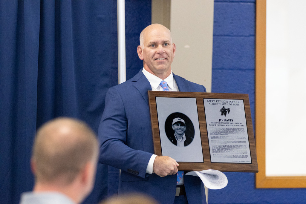 Man holding a framed plaque stands on a stage, smiling. Behind him, blue curtains and a white board.