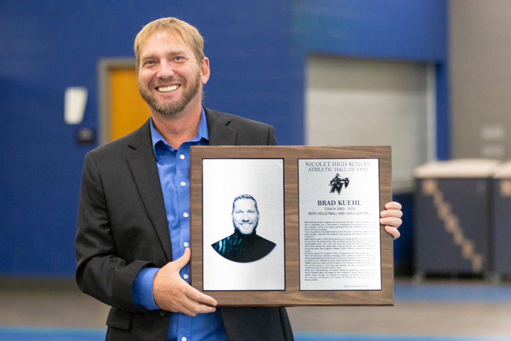 Man in a suit holds plaque with photo and text, smiling, standing in a blue room.