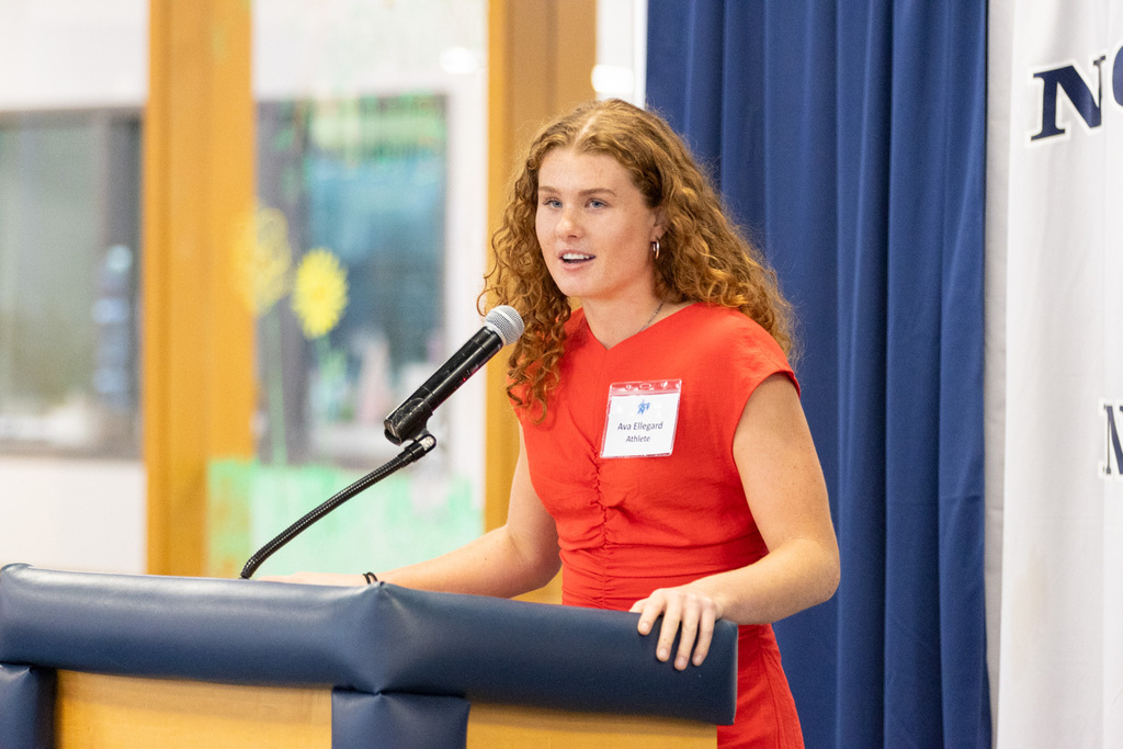 A woman with red hair wearing a red dress stands at a podium, speaking into a microphone.