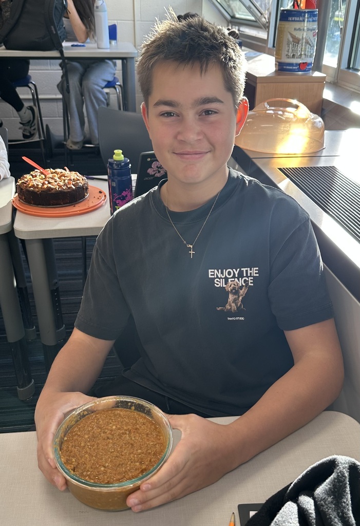 A young person holds a bowl of food and smiles, with a table behind them holding a cake.