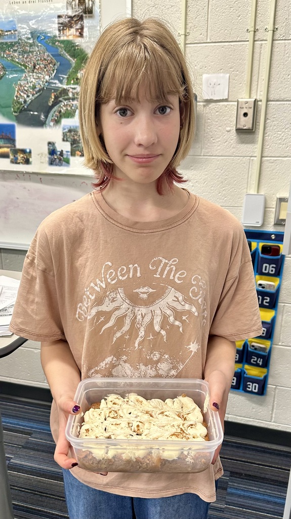 A young person holding a plastic container filled with cinnamon rolls stands in front of a wall with a bulletin board and a map.