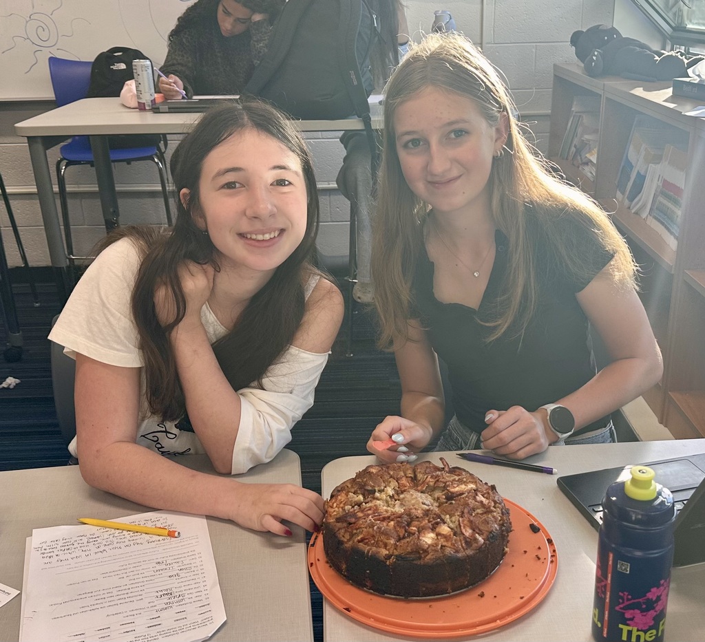 Two women sitting at a desk with a chocolate cake. One holds a pencil and the other holds a knife.