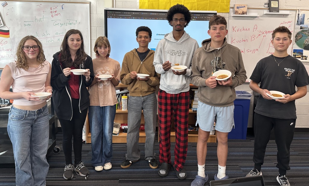 A group of eight students in a classroom with a whiteboard, holding bowls of food.