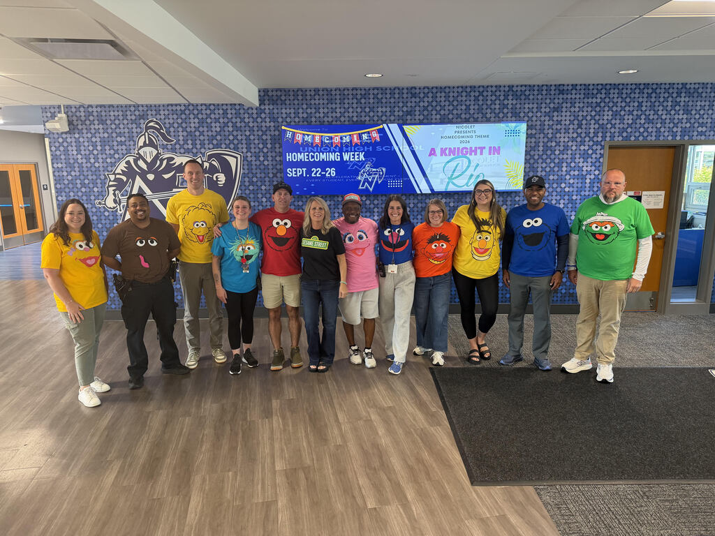 A group of people wearing Sesame Street character shirts standing in a room with blue walls and a banner.