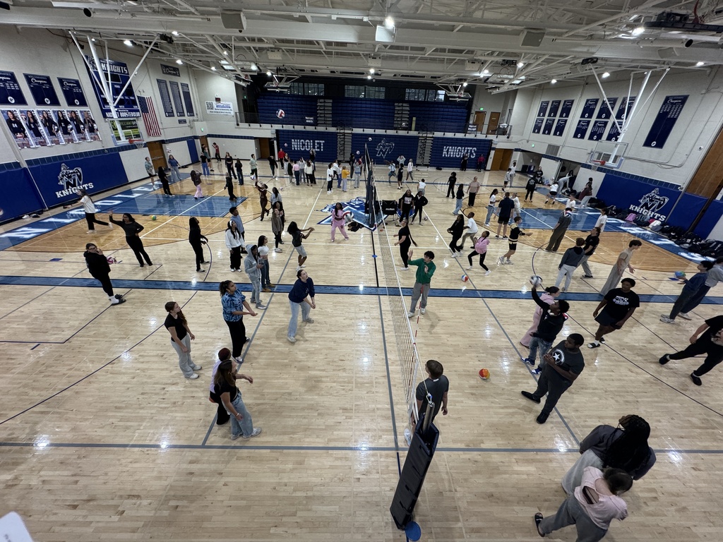 A large indoor basketball court is filled with people playing basketball. The court has white markings, and the walls are blue.