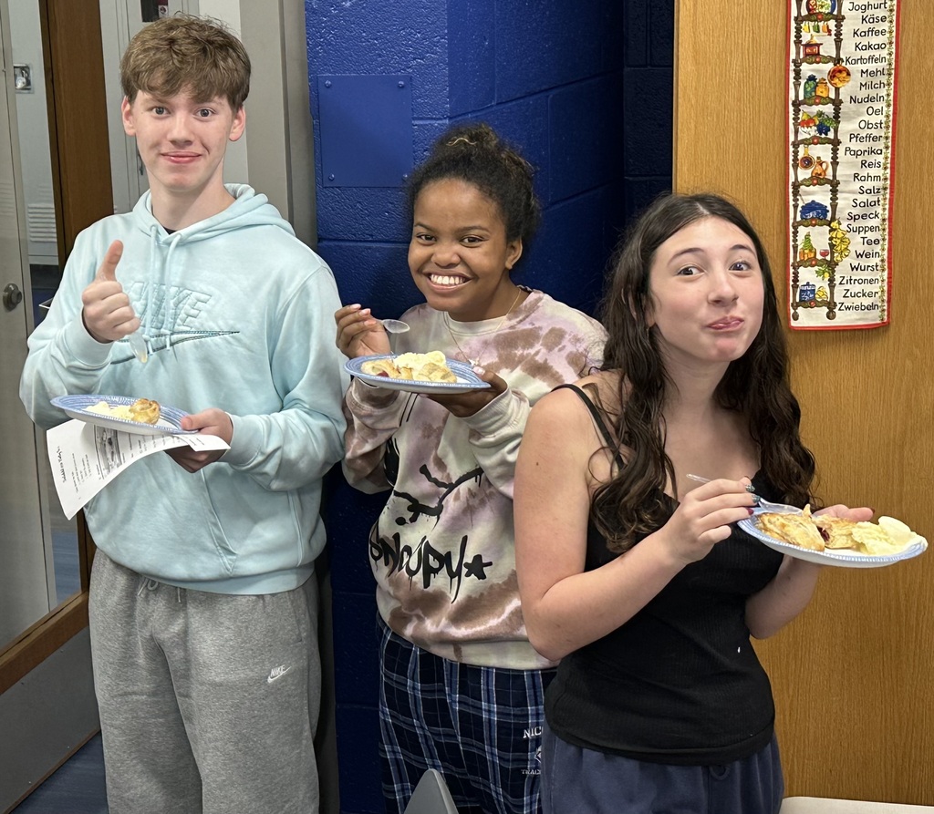Three people stand in a hallway. A male gives a thumbs up. Two females hold plates of food. A bulletin board is on the wall.