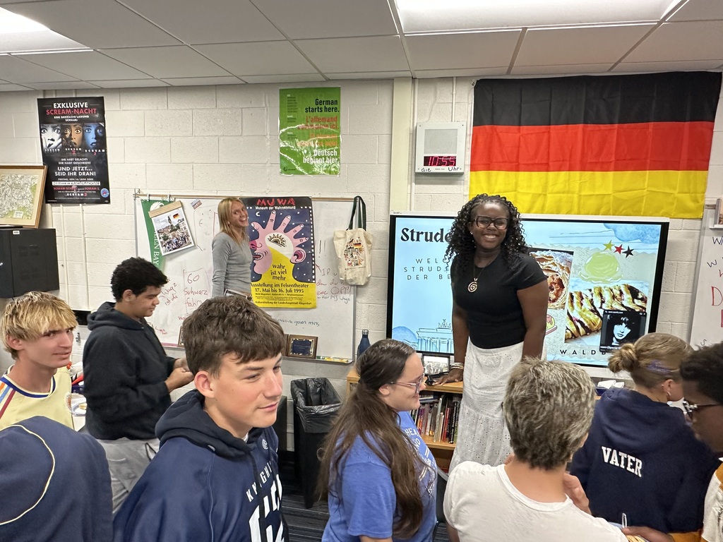 A classroom with students seated and standing. A woman stands in front of a screen. Flags hang on the wall.