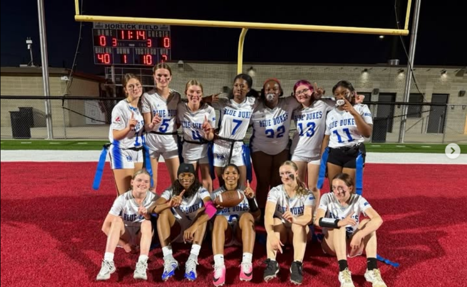Girls' football team, in uniform, posing on a field with a red surface and a football goal.