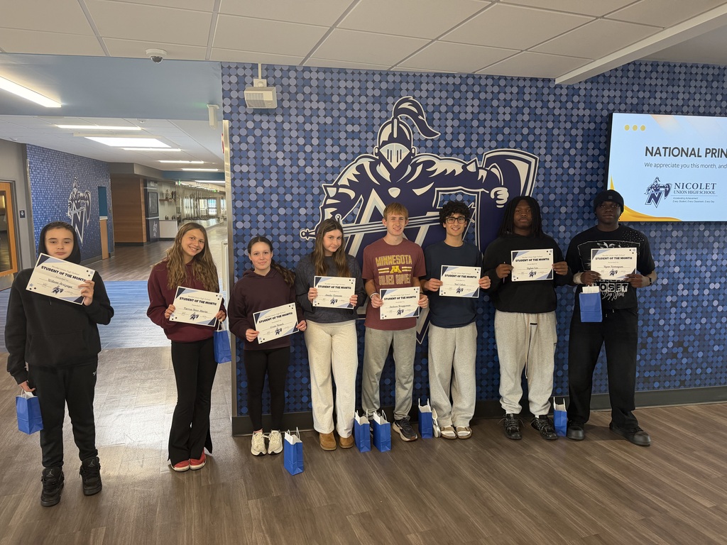 A group of students stand in a corridor, holding certificates. Behind them, a wall features a blue design.