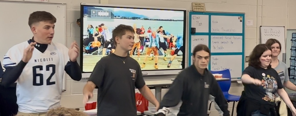Several students in a classroom participate in a performance with a large screen showing a sports game.