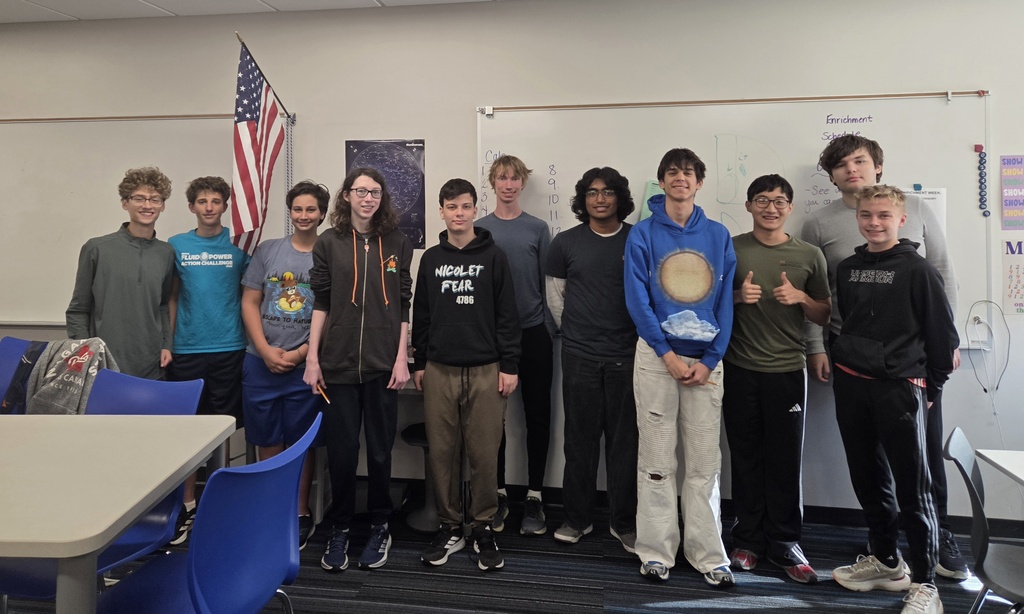 Students in a classroom, lined up with a flag and whiteboard in the background.