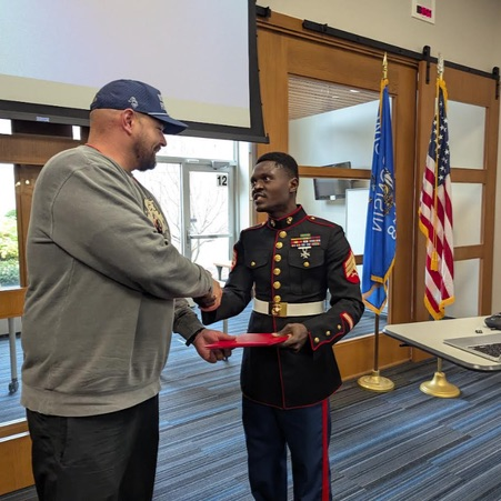 A man in uniform holds a red ribbon and shakes hands with a man in gray.