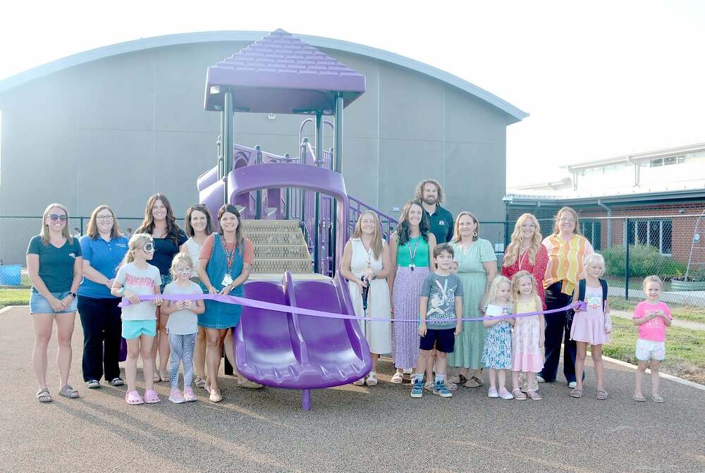 Adults and children gather behind a purple ribbon for a ribbon-cutting ceremony in front of the new purple playground equipment.