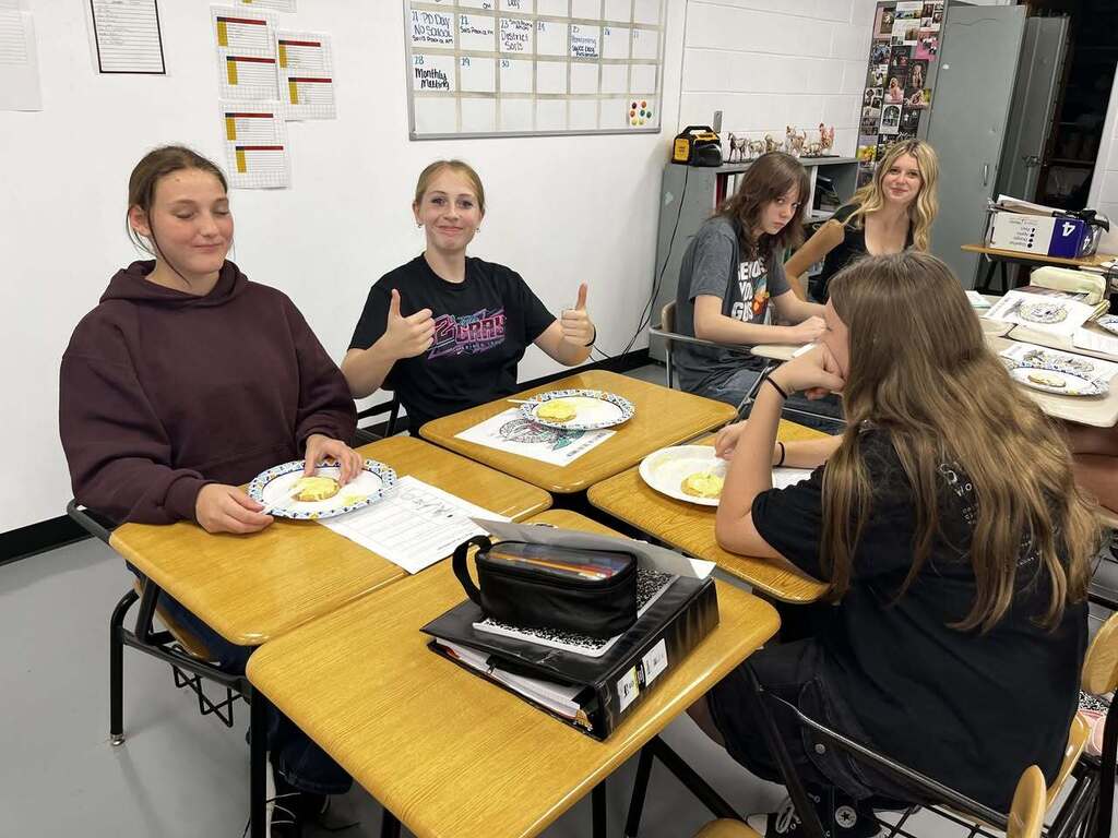 Students at table eating cookies. One student with thumbs up and smiling.