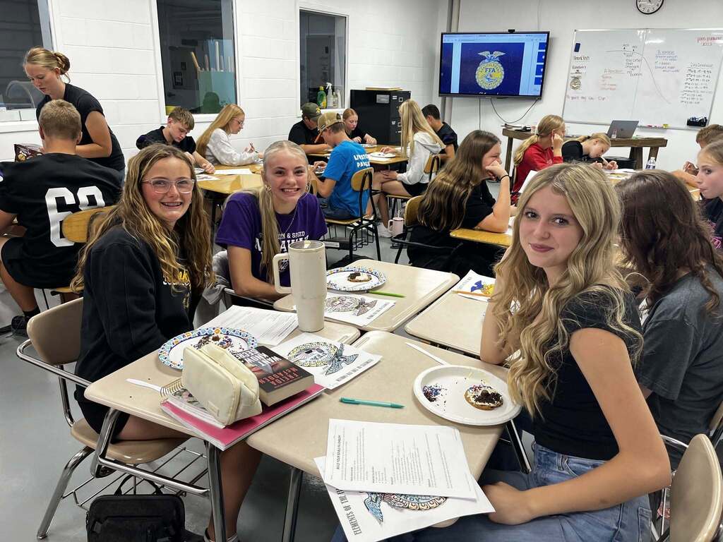 Classroom of students working on assignments and eating cookies. 3 students looking at camera smiling