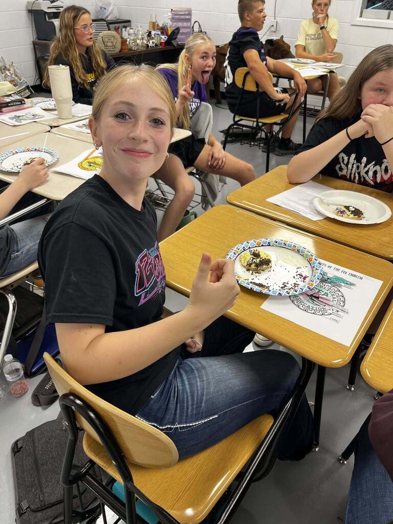 Student looking back at camera while sitting at desk eating a cookie. Student in background making a funny face at the camera