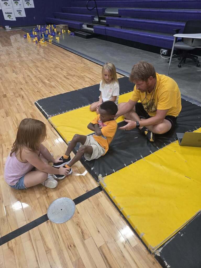 A kindergarten student performs a sit-up while a classmate holds her feet and the physical education teacher monitors the activity.