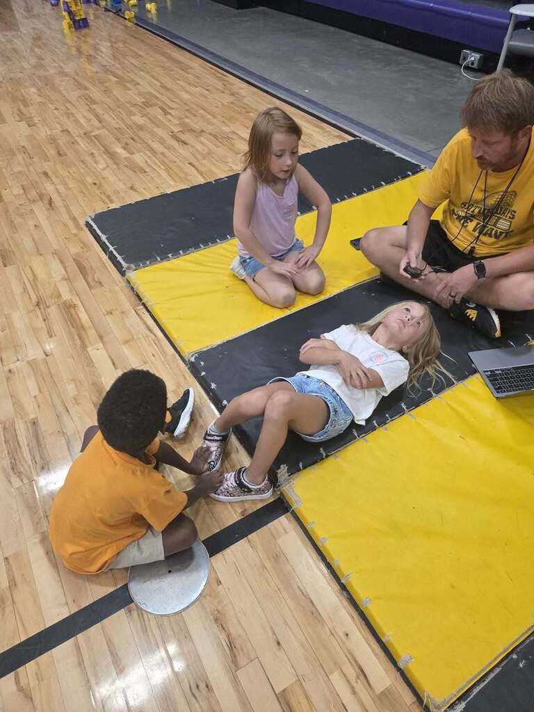 A kindergarten student lies on a gym mat while a classmate holds her feet and the physical education teacher supervises.