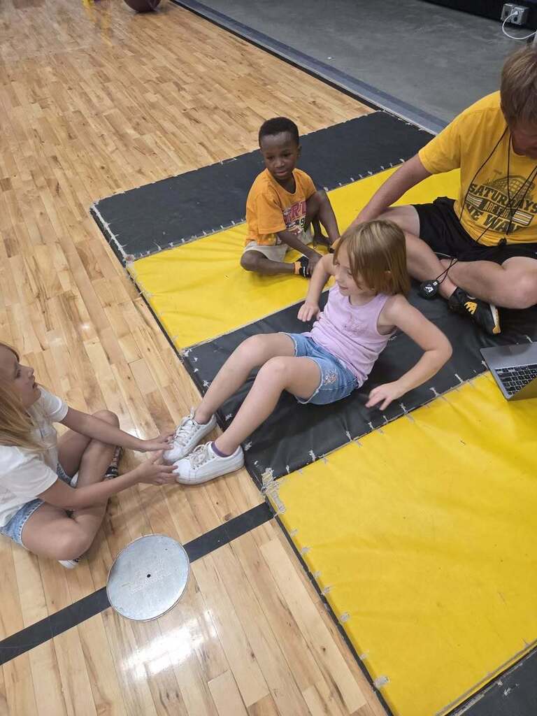 A kindergarten student prepares to perform a sit-up while a classmate holds his feet and the physical education teacher provides instructions.