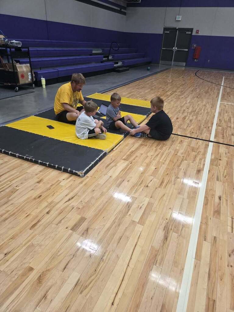 Three kindergarten students sit together on the gym floor near a mat while the physical education teacher leads the activity.