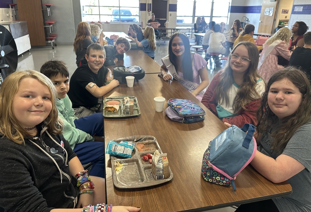 A Homecoming Court member wearing a sash sits with middle school students at a cafeteria lunch table.