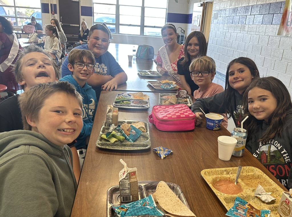 A Homecoming Court member wearing a tiara and sash joins middle school students at their cafeteria table.
