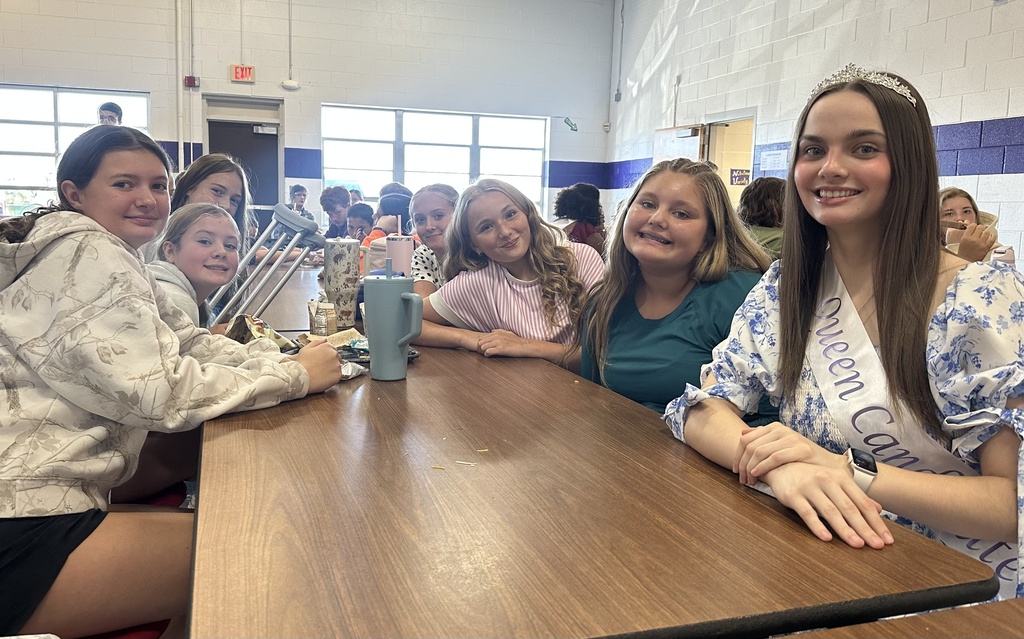 A Homecoming Court member wearing a tiara and sash smiles with middle school students at a lunch table.