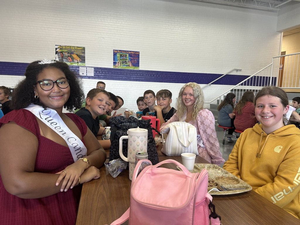 Two Homecoming Court members wearing sashes sit with middle school students during lunch.