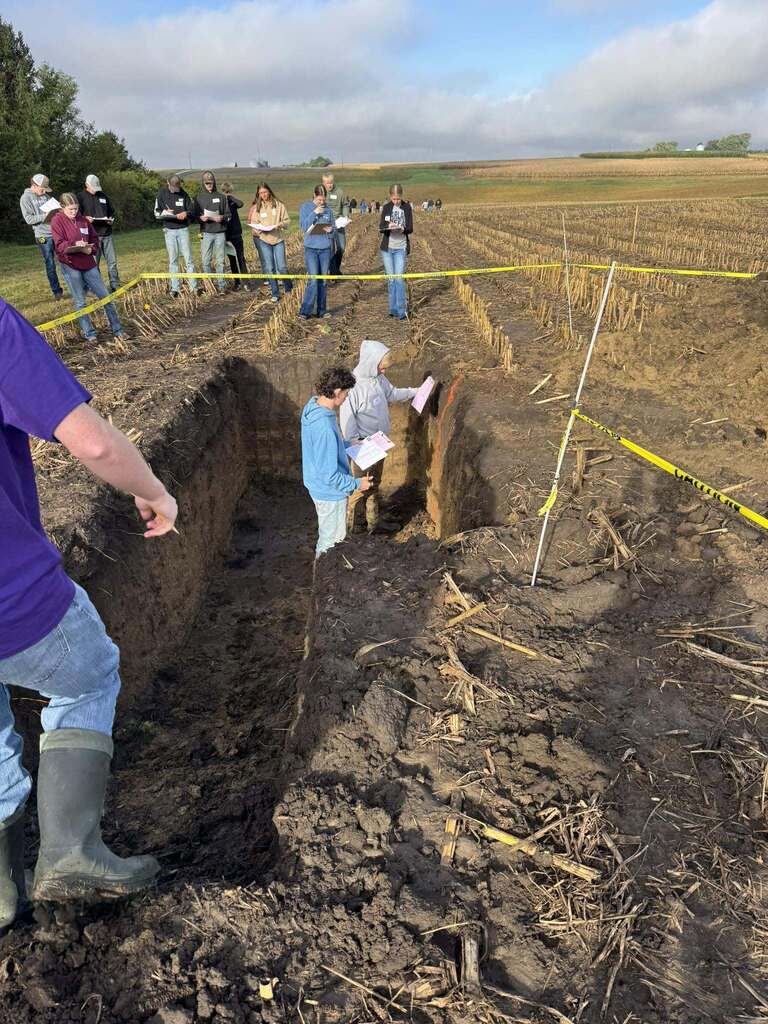 Two students study the wall of a soil pit as a group takes notes behind the caution tape.
