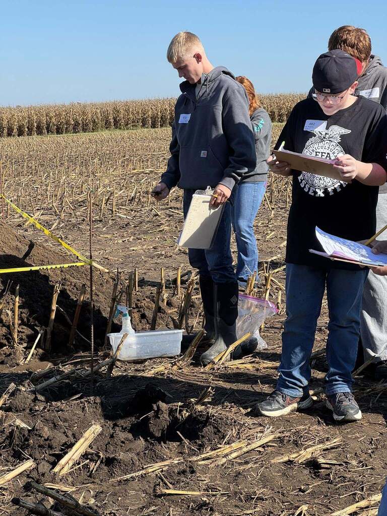 Students examine soil samples and record observations beside a pit in a harvested cornfield.