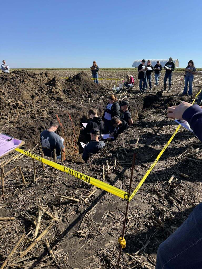 A group of students studies exposed soil layers inside a pit while others take notes nearby.