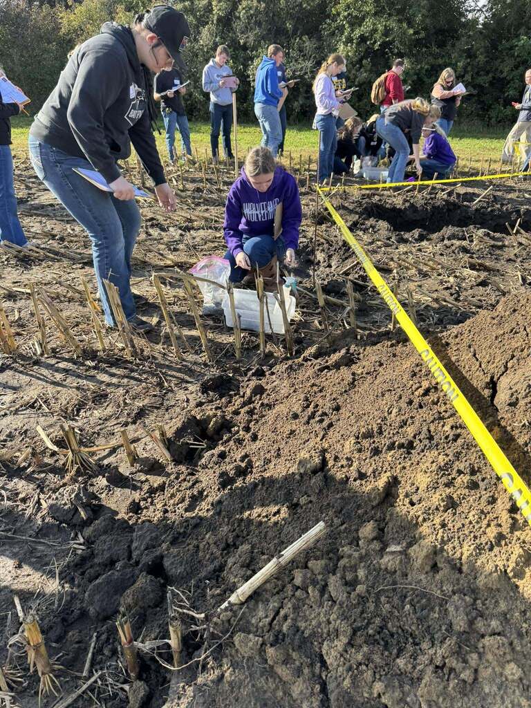 Two students examine soil near a marked pit while other participants work across the field.