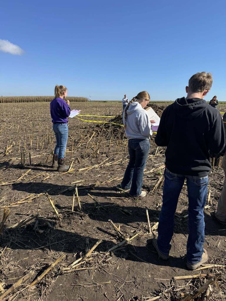 Students read their notes beside a marked soil pit in a harvested field.