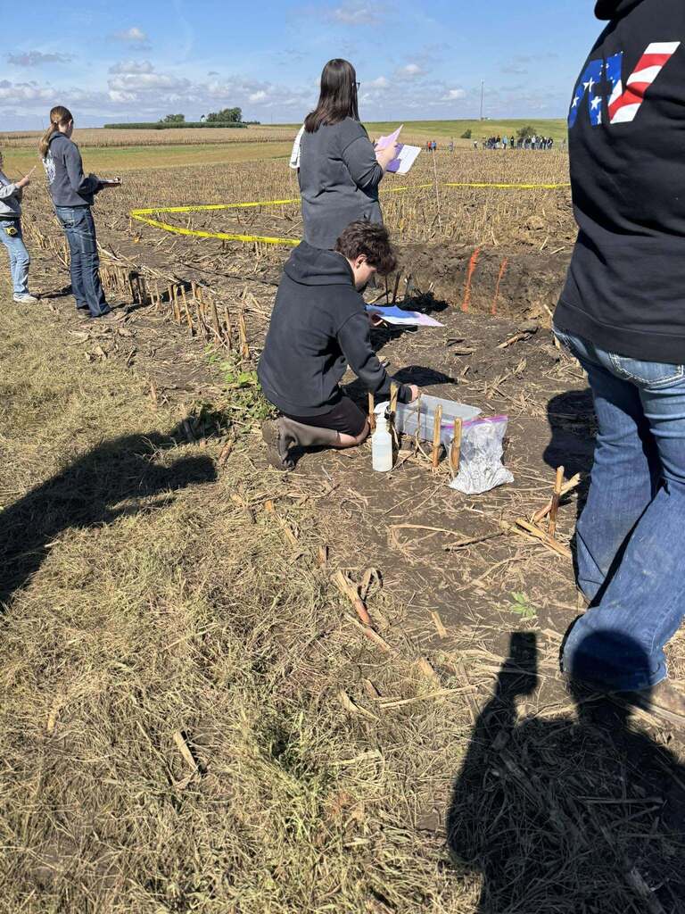 A student kneels beside a soil pit to examine a sample as other participants review their notes.