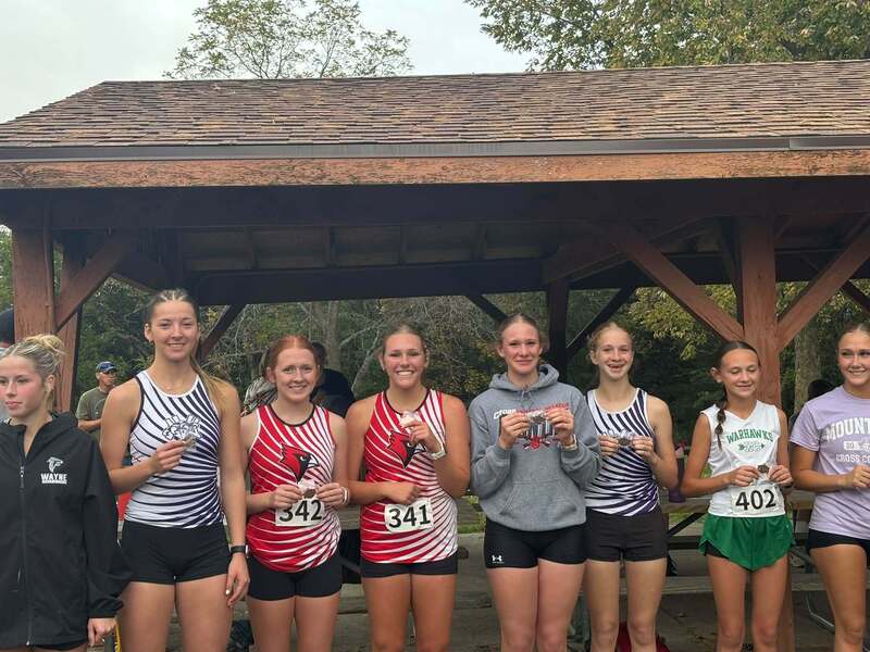 Girls cross country medalists from several schools, including two Nodaway Valley runners, display their medals in front of a park shelter.