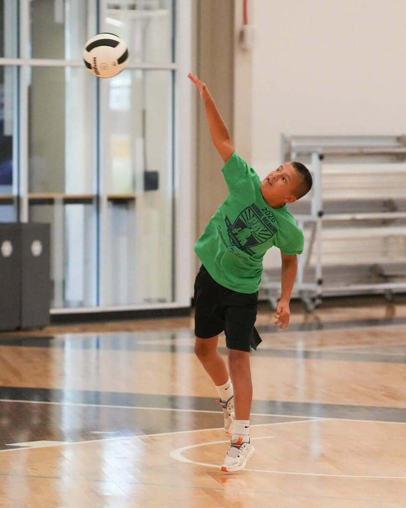 A boy wearing a green shirt and black shorts on a court, reaching up to hit a ball.
