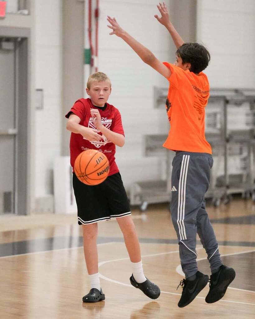 Two boys play basketball in a gym. One boy holds a ball and looks at the other boy who jumps.