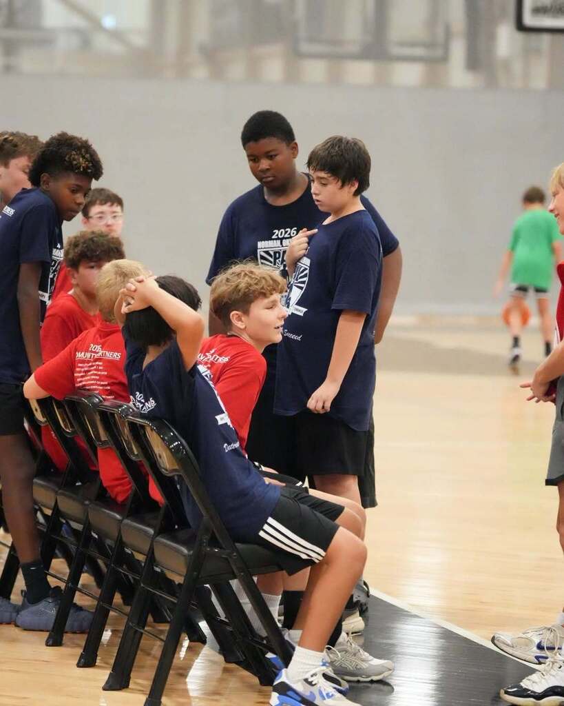 Children seated and standing in chairs at a basketball court, with some focused on a speaker in front of them.