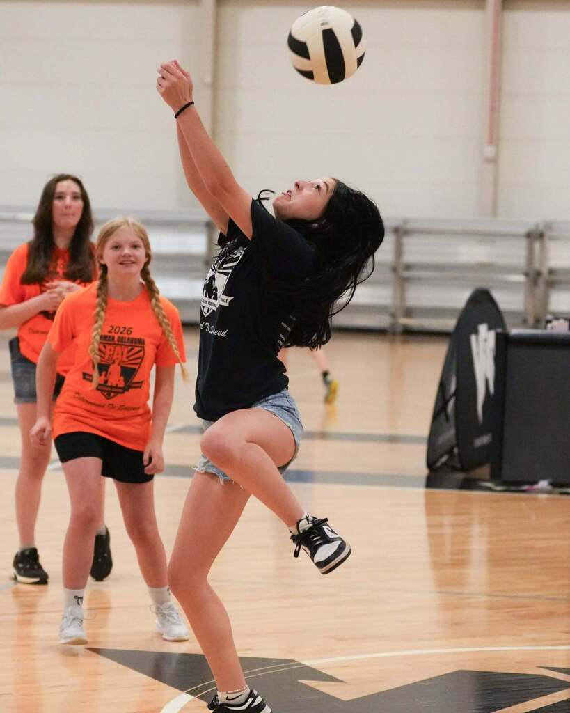 A girl in black shirt and denim shorts jumps and spikes a ball indoors, with two other girls watching.