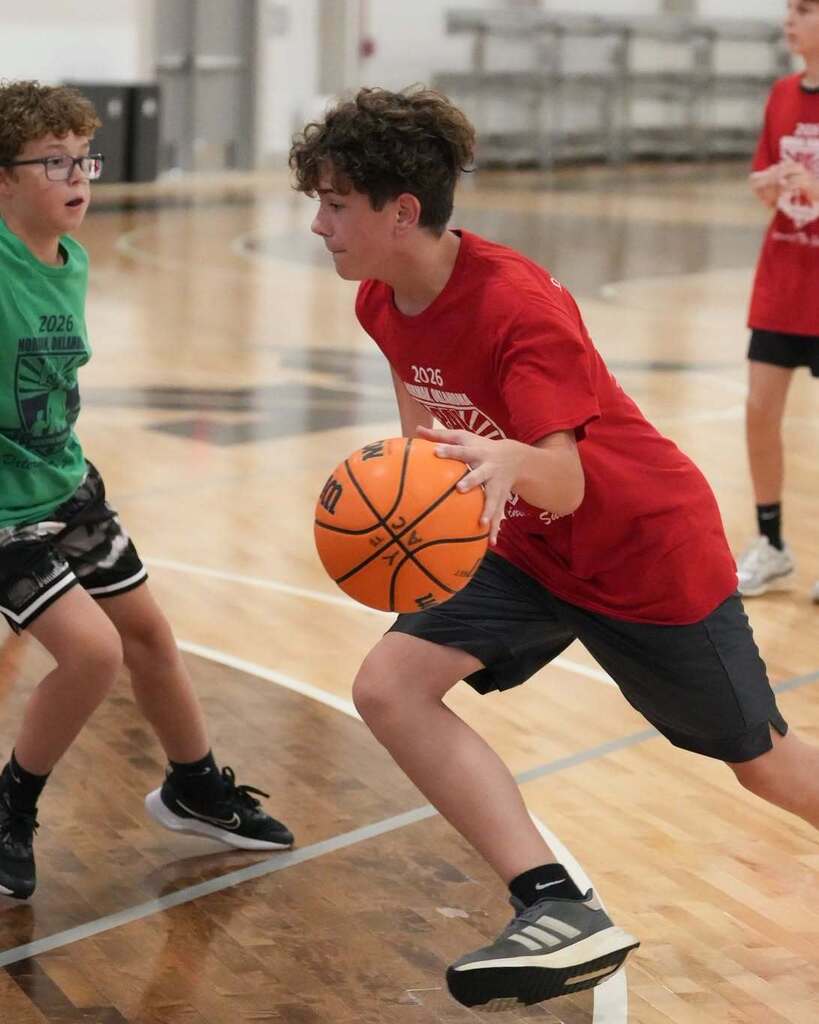 Two kids play basketball indoors, one dribbling the ball and another approaching. A third child stands behind them. The floor has markings.