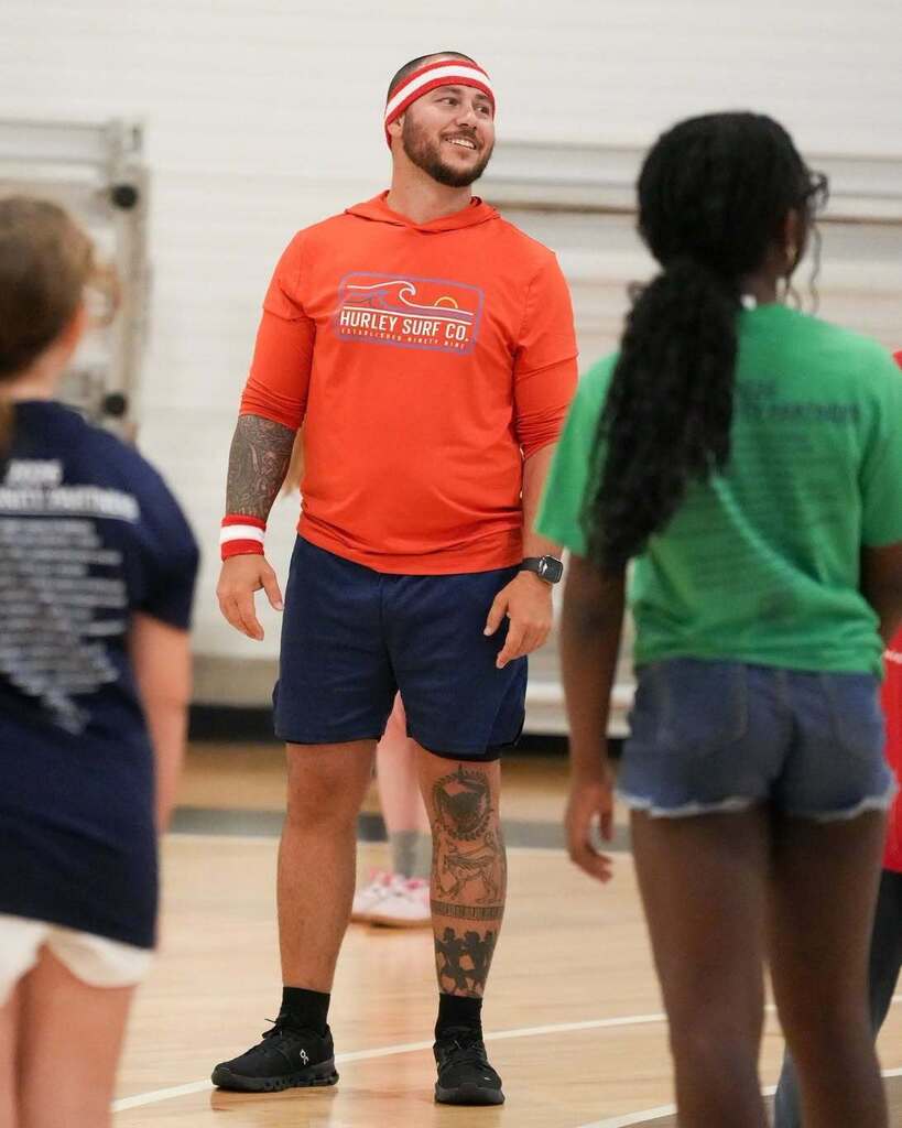 A man in an orange shirt and blue shorts stands in a gymnasium with three girls in colored shirts.