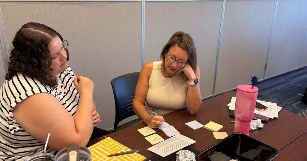 Two people sit at a desk, engaging with cards and sticky notes. One person looks down, the other looks sideways.