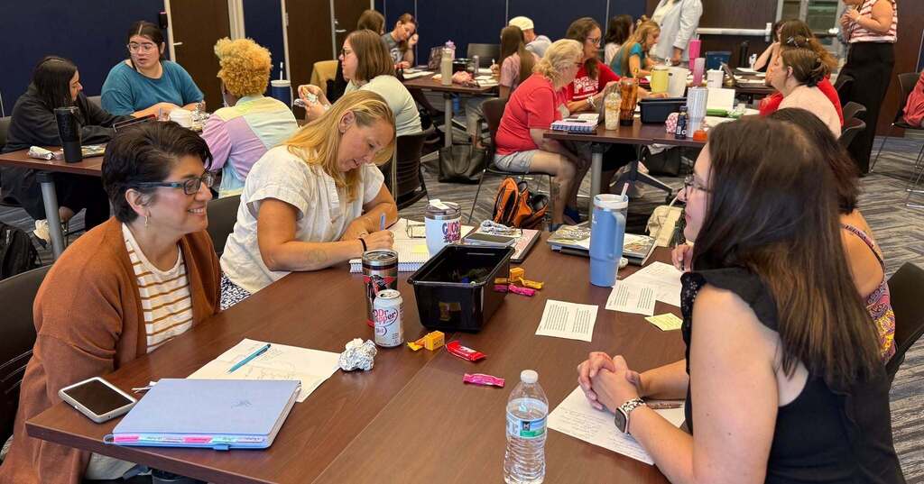 A group of people sit around a table in a meeting room, engaging in discussions, with papers, pens, and water bottles on the table.