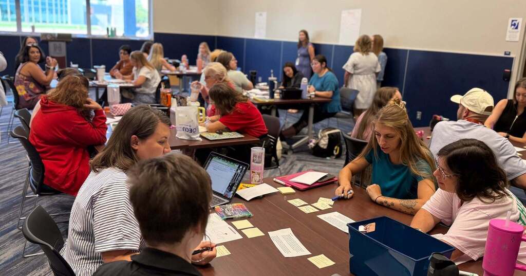 Many people sit around tables with papers, a laptop, and sticky notes in a room.