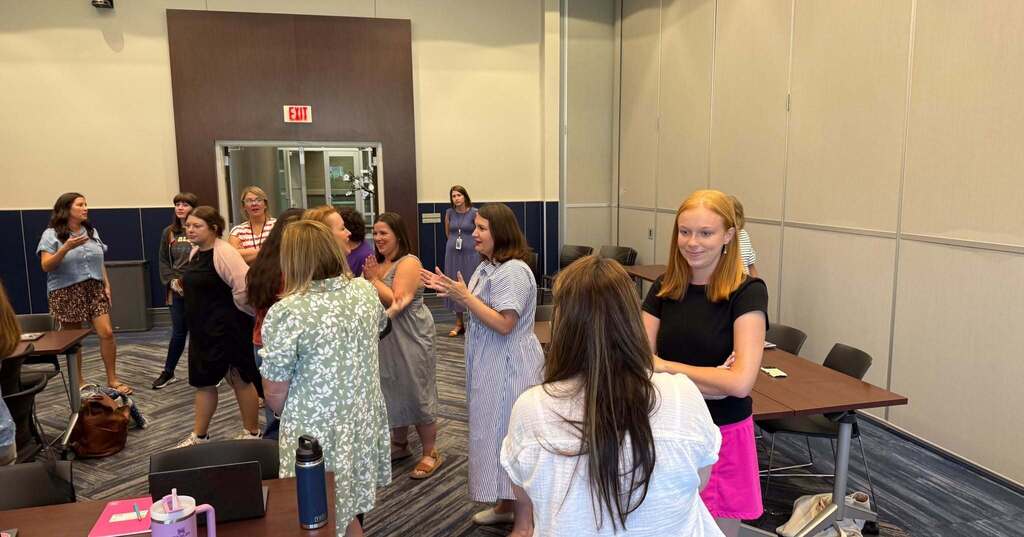A group of women are gathered in a room. Some are standing, others are talking. The room has tables, chairs, and a wall with an exit sign.