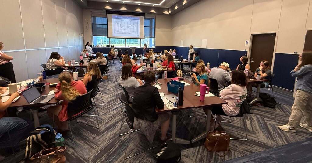 Large group in a meeting room with laptops, one person at a podium. White walls, blue carpet, overhead lights.