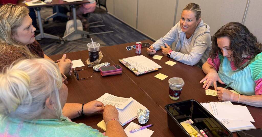 Five women gather around a table, each holding a notepad and pen. Yellow sticky notes are scattered across the table.