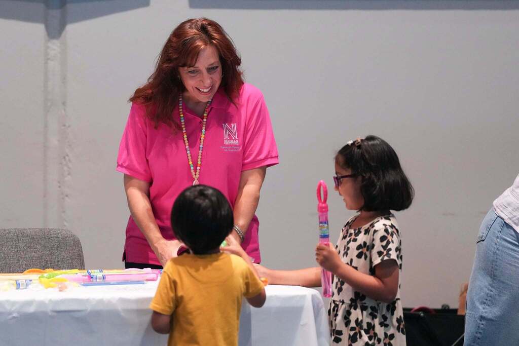 A woman in pink, with two children, one holding a pink object, at a table with a white cloth.