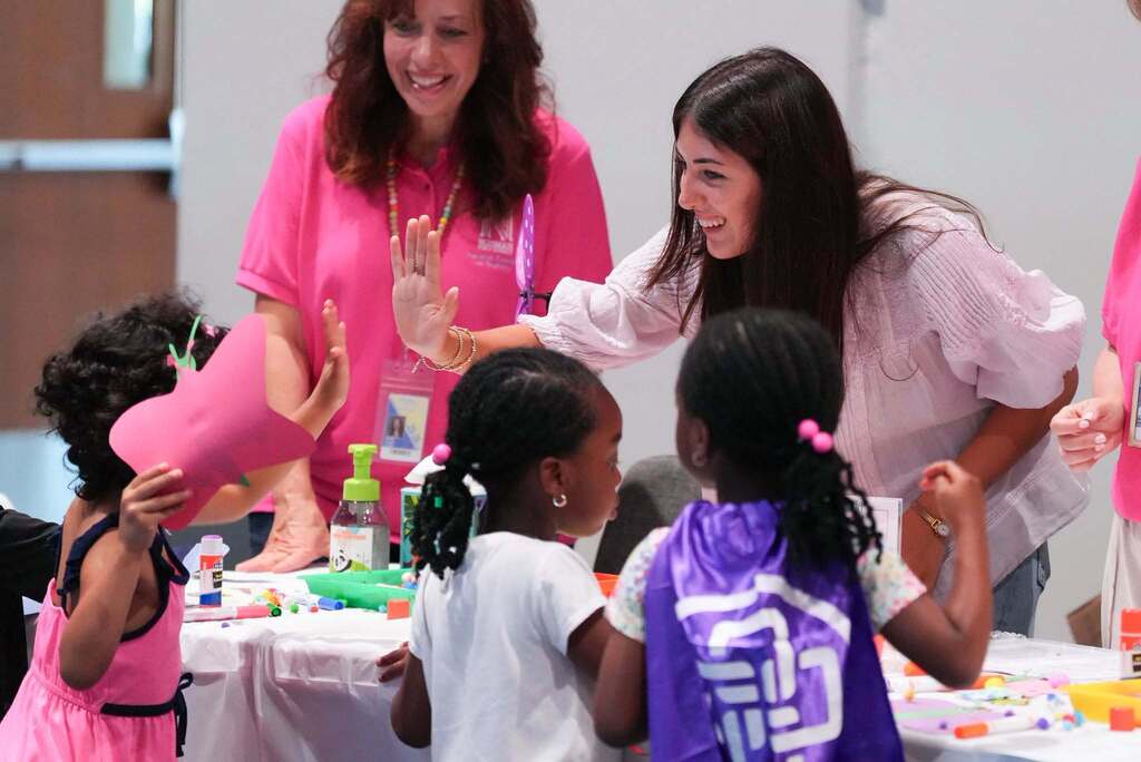 A woman assists children in an indoor activity area, with several children wearing pink dresses and one wearing a purple cape.